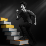Black-and-white photograph of a young man smiling confidently while climbing a staircase made of colorful books. The books are the only elements in color, featuring vibrant reds, yellows, greens, and blues, set against a dark, dramatic background with a spotlight highlighting the scene.