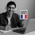 A young man studying French, smiling at the camera while wearing headphones, seated at a desk with a laptop and notepad. The black-and-white photo highlights a colorful French flag on the notepad.
