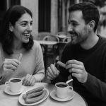 A black and white photo of a man and a woman sitting at an outdoor café table, smiling and talking while eating churros and drinking hot chocolate, with empty chairs and a quiet street in the background. They are able to communicate because they use language learning app.