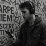 Black and white photo of a young man wearing headphones, leaning against a graffiti-covered wall with large Latin words 'CARPE DIEM' and 'DISCERE LATINUM' painted on it. He is looking at a glowing smartphone screen, symbolizing the modern way of learning Latin with the help of language learning app.