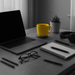 Black and white photograph of a minimalist desk setup with an open laptop, notebook, pens, glasses, and headphones. The only colored element is a yellow mug placed next to a small potted plant, standing out against the monochrome scene.