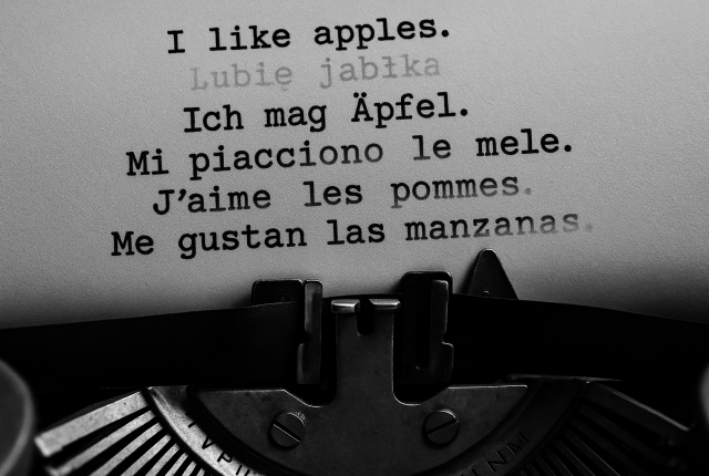 Black and white 16:9 close-up photograph of a typewriter merging with a modern keyboard, with fragments of sentences in English, Polish, German, Italian, French, and Spanish appearing and fading across the page and screen. The image symbolizes how Taalhammer transforms traditional language learning into adaptive, AI-driven full-sentence fluency.