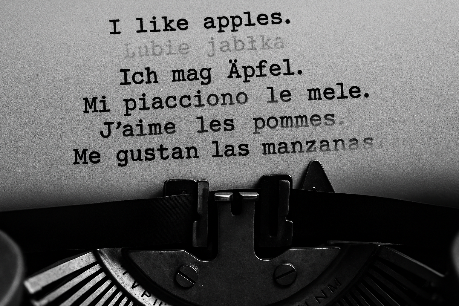 Black and white 16:9 close-up photograph of a typewriter merging with a modern keyboard, with fragments of sentences in English, Polish, German, Italian, French, and Spanish appearing and fading across the page and screen. The image symbolizes how Taalhammer transforms traditional language learning into adaptive, AI-driven full-sentence fluency.