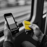A grayscale photo taken from a first-person perspective, showing someone sitting by a train window while studying. They hold a smartphone displaying a flashcard app in one hand and a small stack of physical flashcards in the other. One flashcard is highlighted in yellow with the word “taalhammer” written on it.