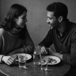 Two people sitting close together at a small table, sharing a quiet meal and leaning in toward each other in warm conversation. The scene is black and white, with a calm, intimate atmosphere. They are able to communicate because they use a language learning app.