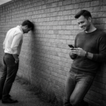 “Black-and-white 16×9 photo showing two people at a wall: one leaning forward with their head against the wall in frustration, the other relaxed, smiling at a phone — a visual metaphor for a language learning app that helps learners move past plateaus.