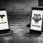 Black-and-white, 16:9 ultrarealistic photo showing two smartphones on a wooden desk, facing each other. One phone displays the minimalist Taalhammer logo (black “T” shape with a small square beneath), the other shows the Duolingo owl icon. The scene suggests a comparison between language learning apps, with a clean, editorial, text-free composition.