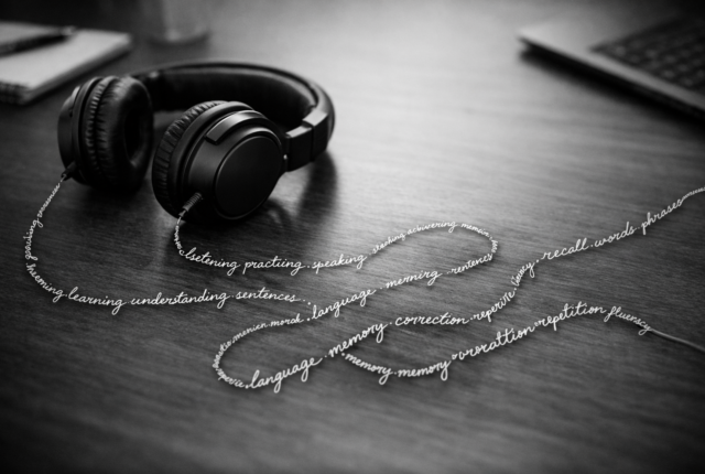 Black-and-white 16:9 photo of over-ear headphones on a desk, with the cable formed from cursive words, symbolising listening to your own sentences in language learning.