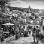 Black-and-white ultrarealistic 16:9 photo of a coastal Portuguese village with whitewashed houses and tiled roofs, a cobblestone street leading downhill to the sea, and people walking and sitting at a small café terrace.