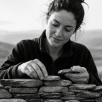 Black-and-white ultrarealistic image of a person outdoors carefully stacking small stone pieces into a structured form, symbolizing active language construction and building fluency through a structured learning system.
