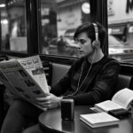 Black-and-white image of a person in a café reading a foreign-language newspaper while listening to audio through headphones, illustrating how everyday activities can become part of a language immersion system without living abroad.