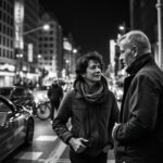 **Alt text:** Black-and-white image of a traveler asking a local for directions on a busy city street at night, illustrating the challenge of accessing language under real-world pressure.