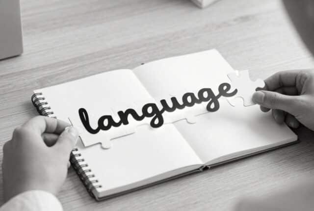Black-and-white, close-up scene of hands assembling puzzle pieces that form the word “language” above an open notebook on a desk, symbolizing building language structure piece by piece.