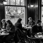 Czarno-biała, niezwykle realistyczna scena z wnętrza przytulnej amsterdamskiej kawiarni nad kanałem. Kilka osób siedzi przy małych okrągłych stolikach pod dużymi oknami, cicho czytając książki i popijając kawę. Przez szyby widać kanał, rowery i wąskie holenderskie budynki, co tworzy spokojną, sprzyjającą refleksji atmosferę.